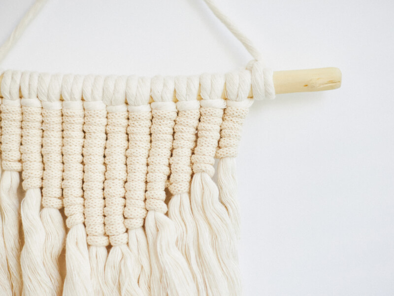 A cream coloured macrame hanging on a pale wooden pole hanger with frayed macrame ends